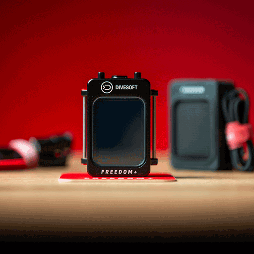 A Divesoft Freedom+ Full Trimix Dive Computer with a black front panel, displayed on a red surface with diving equipment in the background.