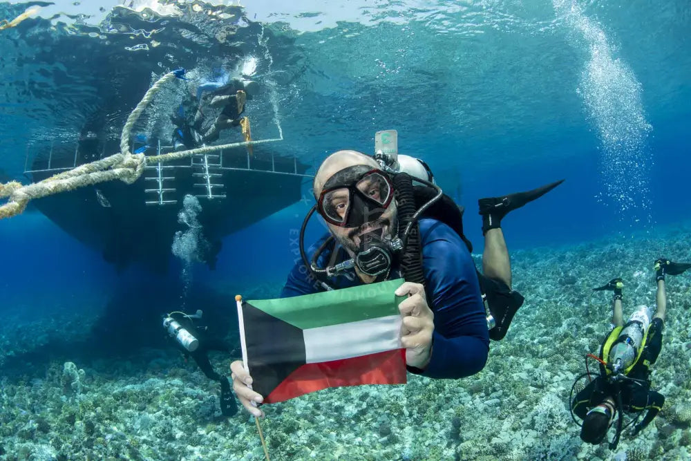 A scuba diver in a blue wetsuit holds the Kuwaiti flag during the full-day Kuwait islands dive trip to Qaruh and Umm Al Maradim