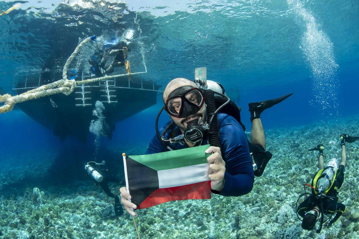 A scuba diver in a blue wetsuit holds the Kuwaiti flag during the full-day Kuwait islands dive trip to Qaruh and Umm Al Maradim