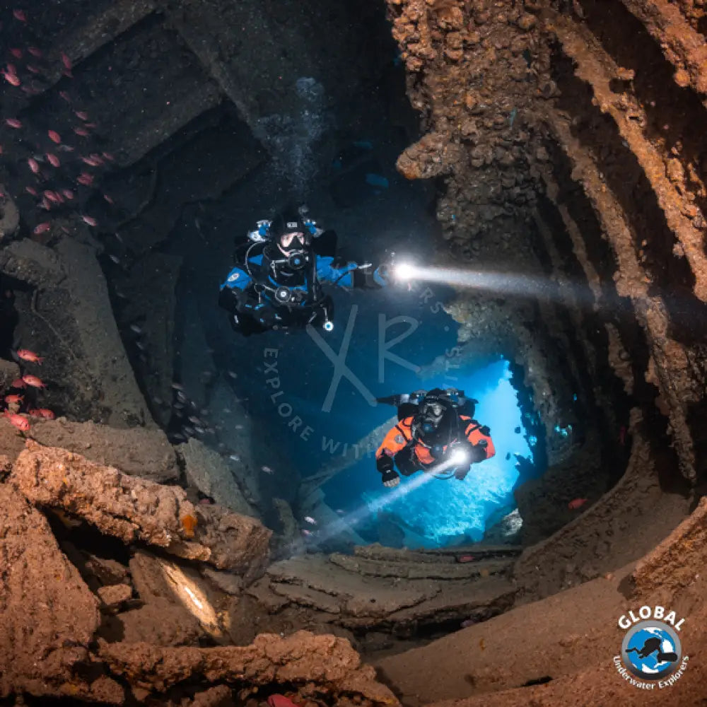 Two GUE scuba divers exploring a wreck with lights on, surrounded by rocky walls and water.
