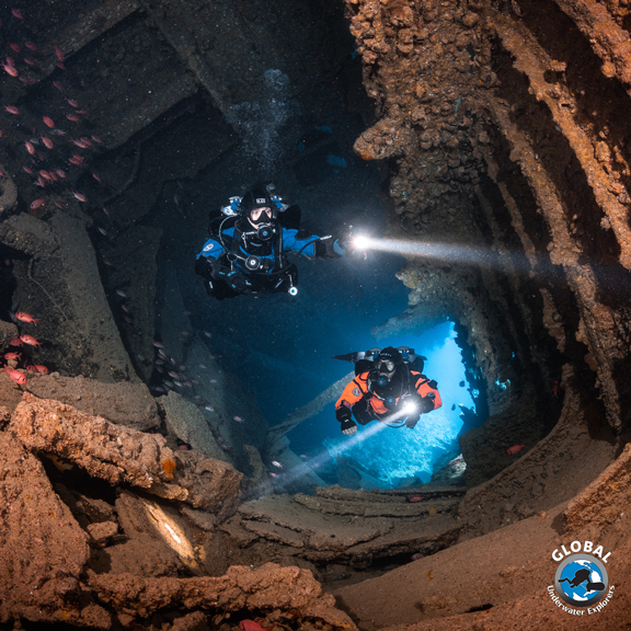 Two GUE scuba divers exploring a wreck with lights on, surrounded by rocky walls and water.