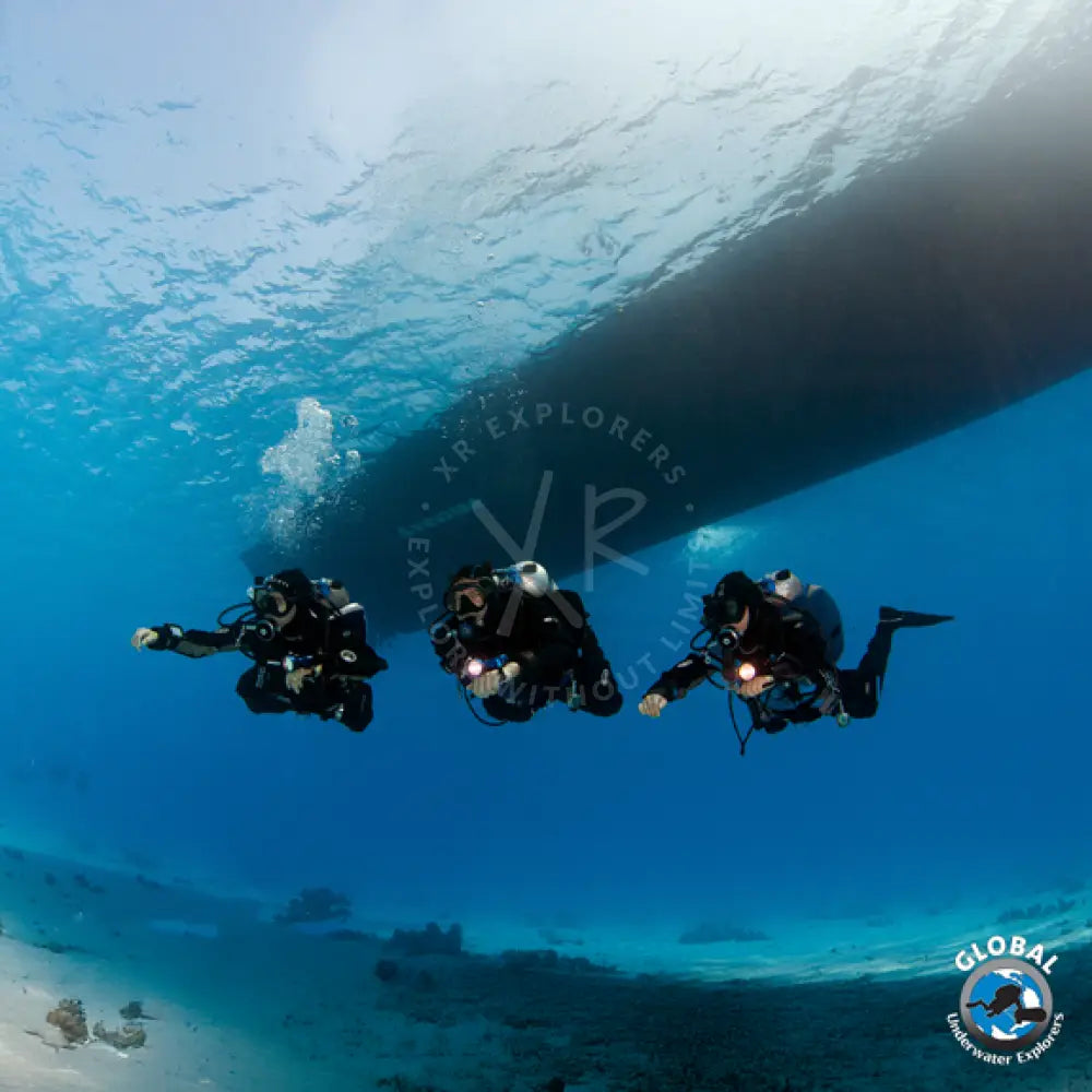 Three GUE basic fundamentals divers underwater with a large shipwreck in the background, featuring the Global Explorer logo.
