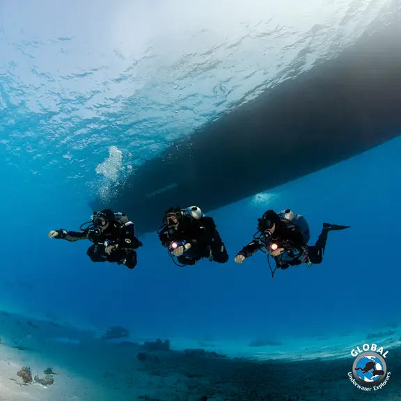 Three GUE basic fundamentals divers underwater with a large shipwreck in the background, featuring the Global Explorer logo.
