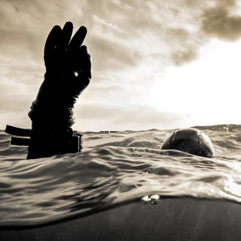 Person in wet suit reaching out from water towards the sky.